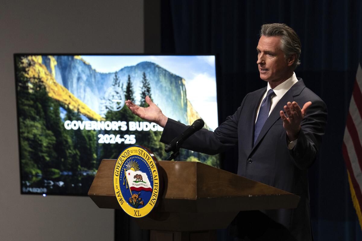 California Gov. Gavin Newsom opens his arms as he speaks from a lectern, a screen at left showing the opening of his budget
