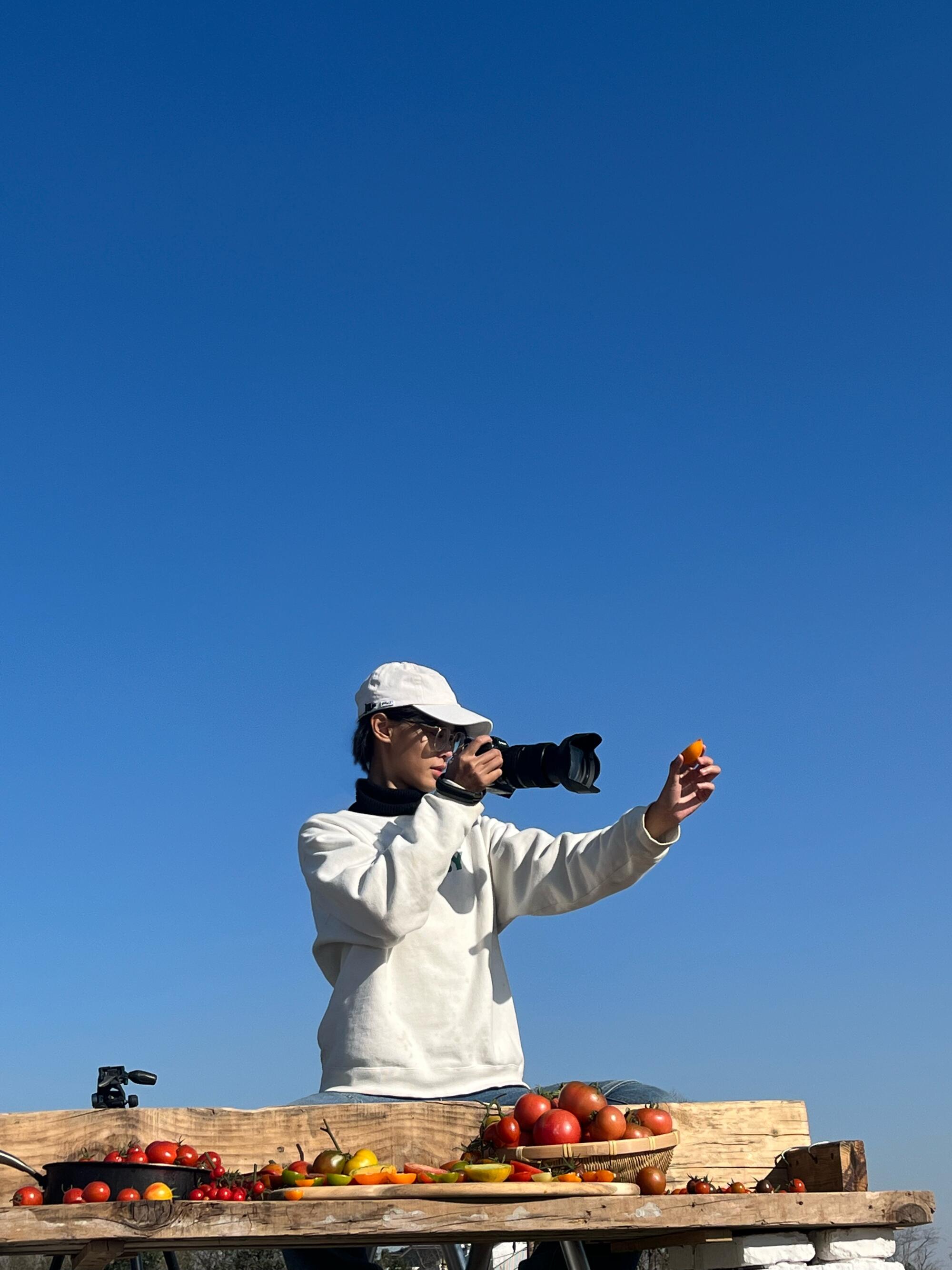 A person in a white sweatshirt and cap holds a camera with long lenses, pointing at a tomato held in the other hand.