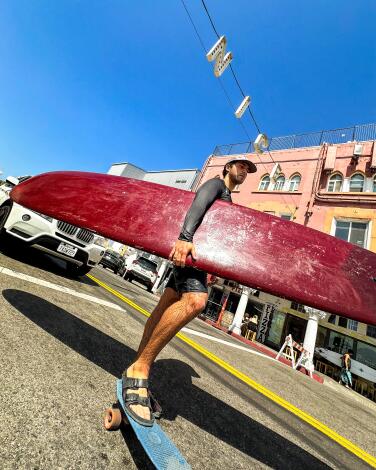 A person stands under the Venice sign holding a surfboard, one foot resting on a skateboard