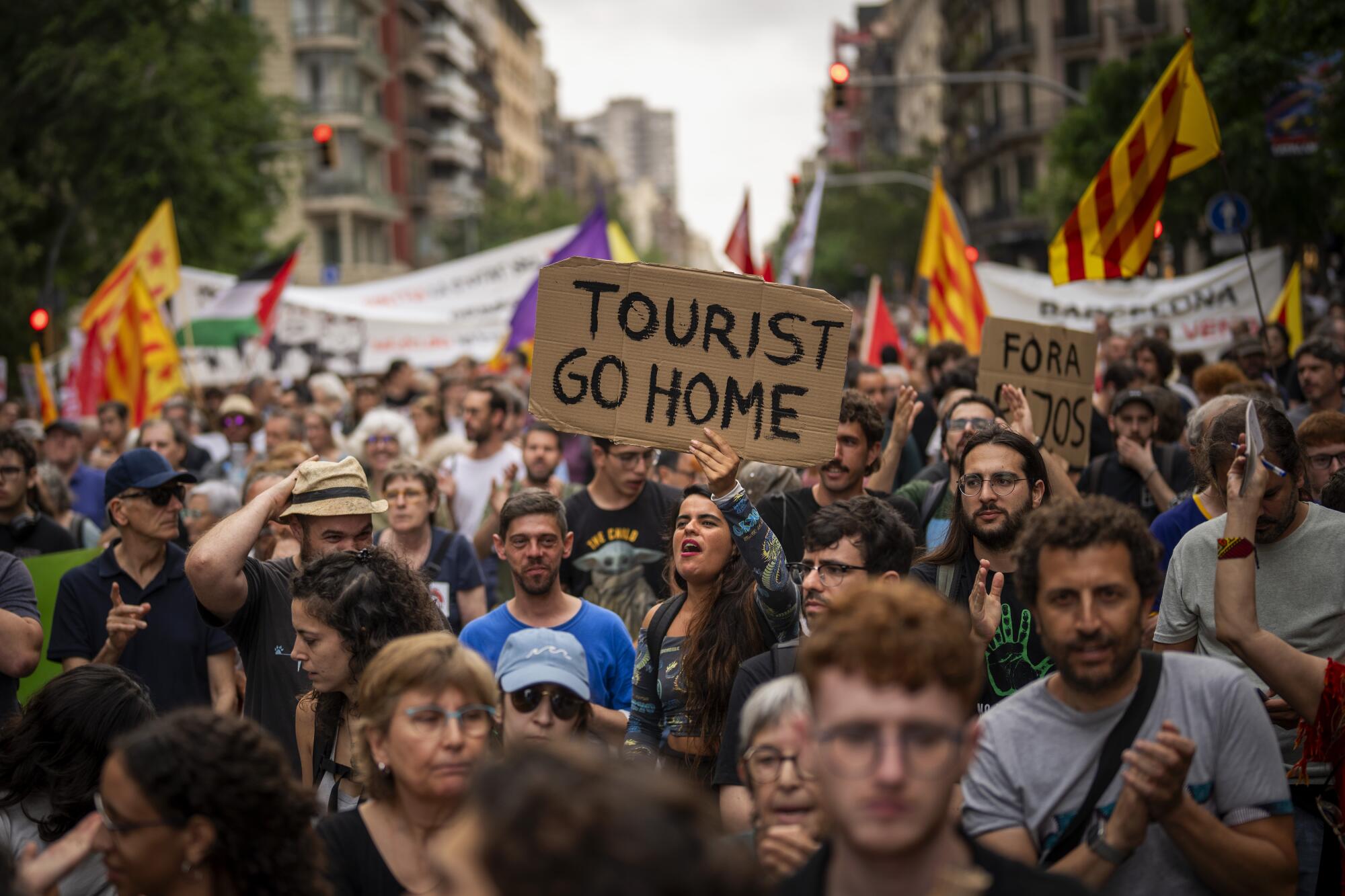 A crowd of protesters marching on a city street, many hoisting banners and one with a homemade sign reading "Tourist go home"