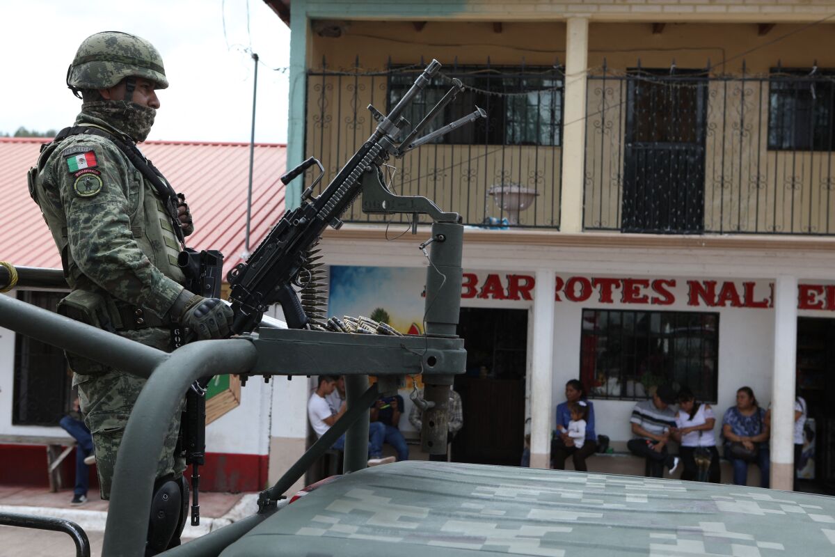 A soldier stands behind a mounted weapon in the back of a military vehicle in front of a building