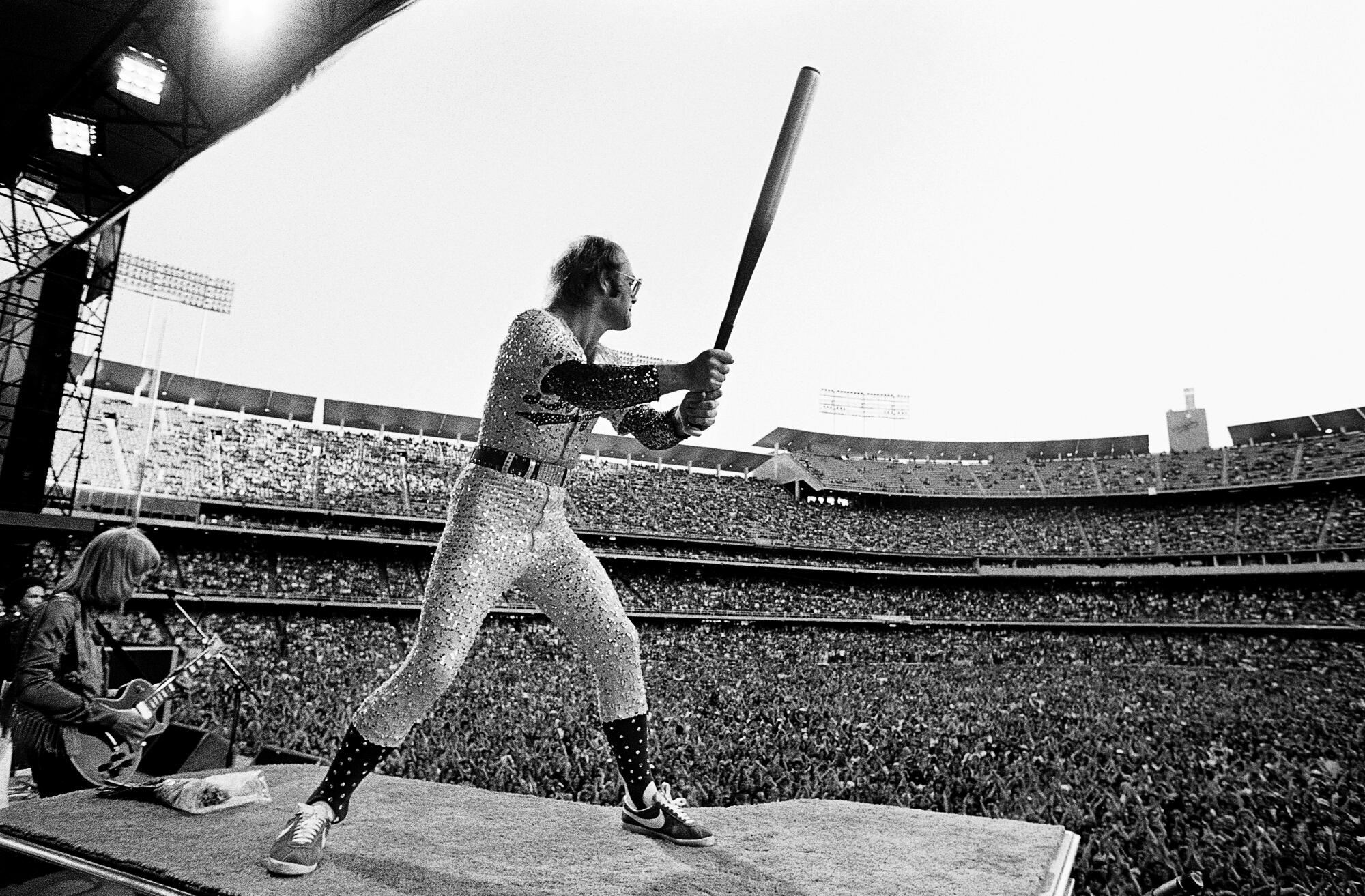 A rock musician stands atop a piano wielding a baseball bat in front of a stadium full of fans.