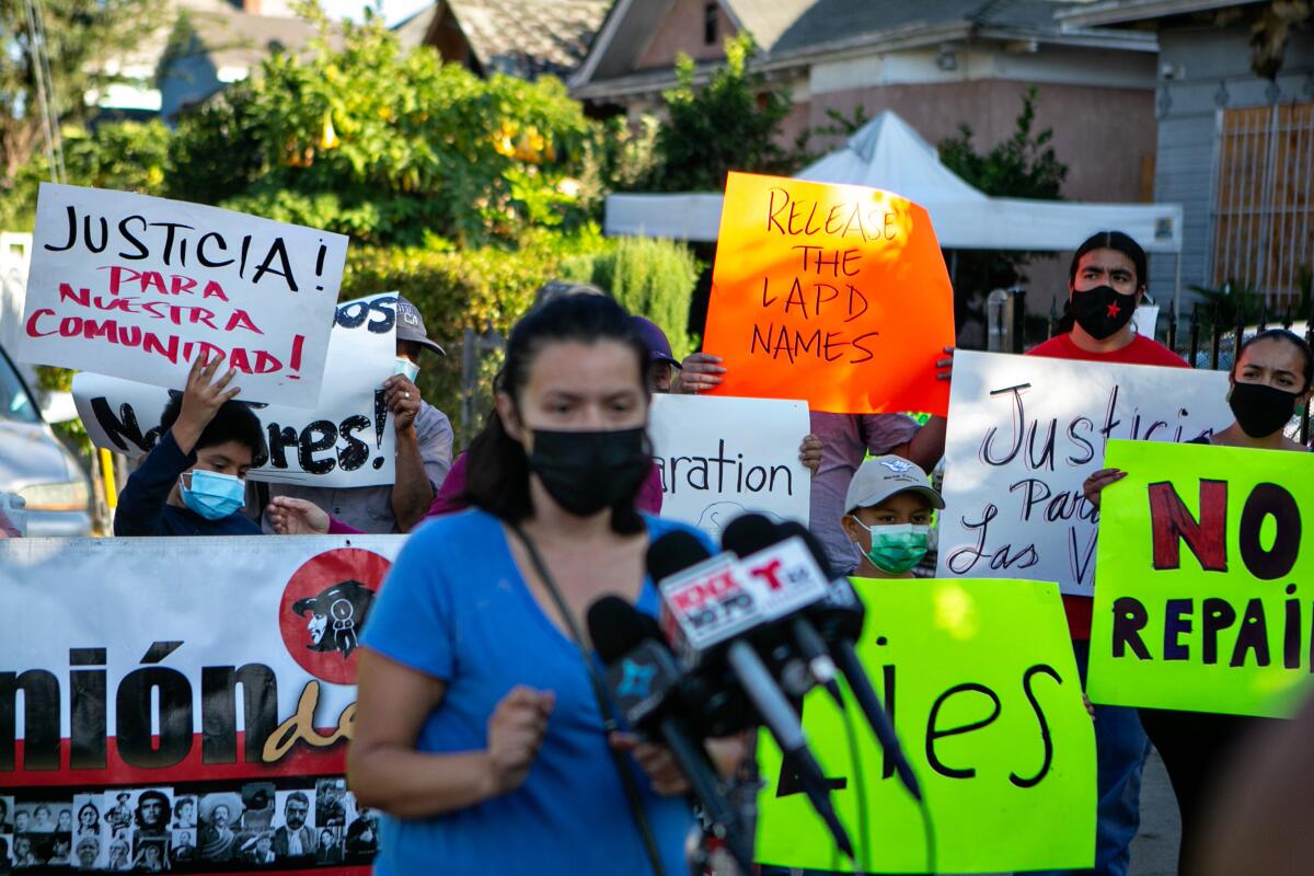 A person wearing a face mask speaks into microphones while people behind them hold signs.