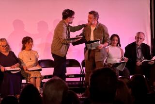 Two men holding scripts perform their parts of a play while other seated actors onstage look on.  