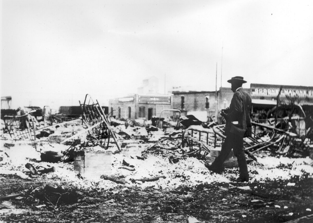 A black-and-white photo of a man in a hat and jacket walking toward the remains of iron beds lying amid debris