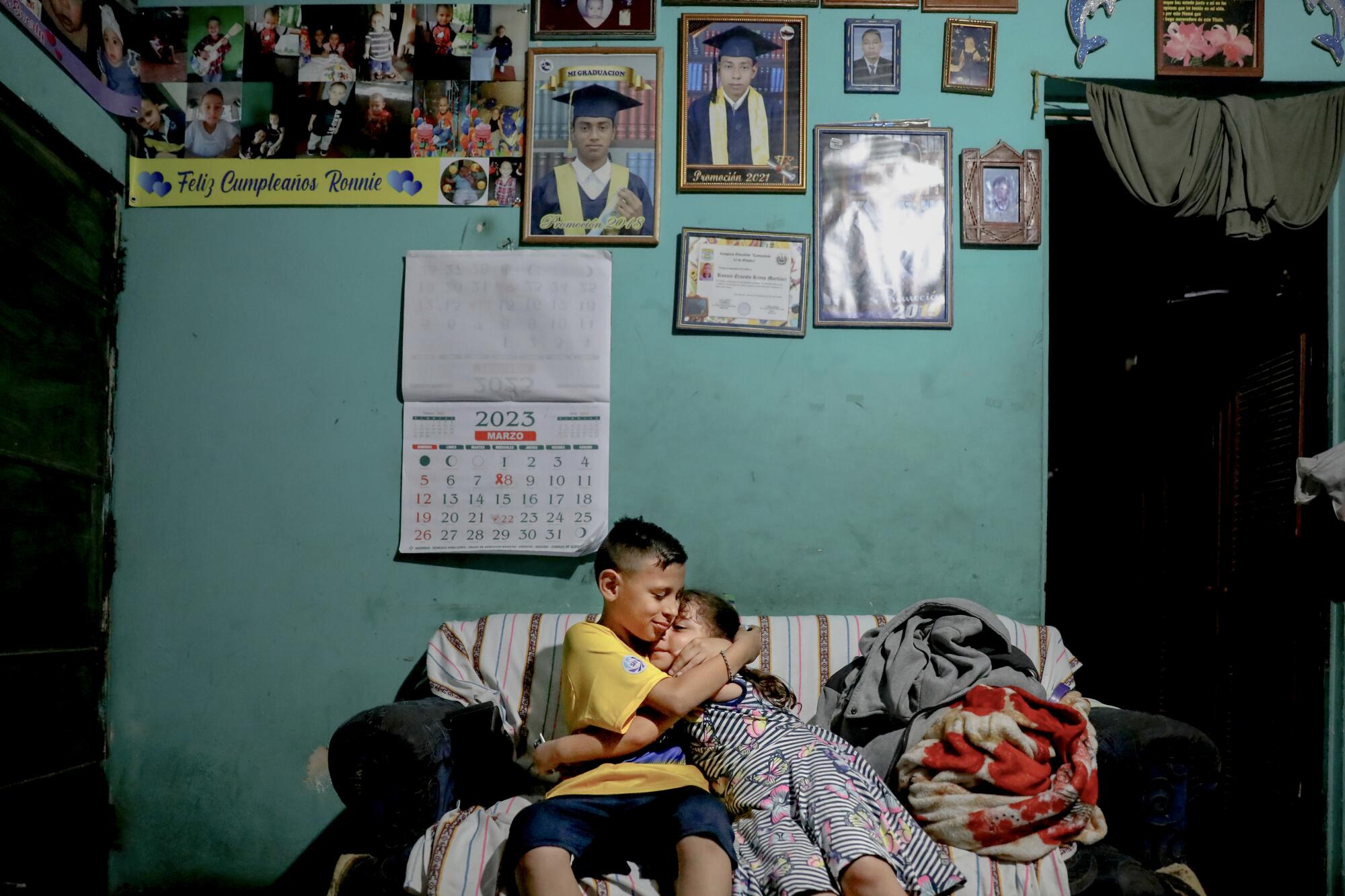 A boy and a girl embrace on a couch in front of a turquoise-colored wall adorned with pictures 