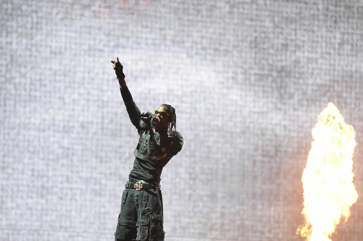 A distant shot of Travis Scott onstage next to a flame, holding his right hand up with a microphone held to his mouth