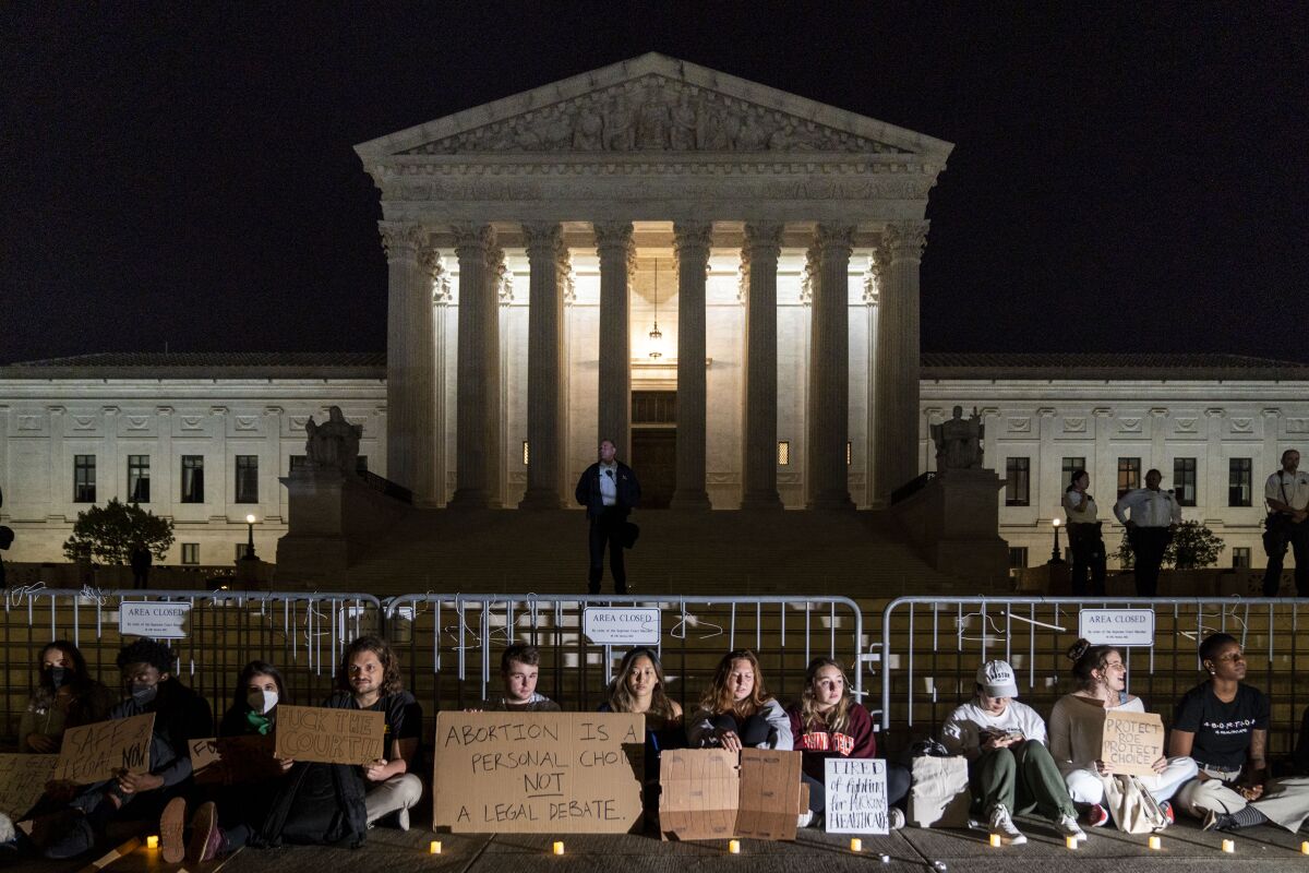 Demonstrators with cardboard signs sit along a barrier at night in front of the Supreme Court