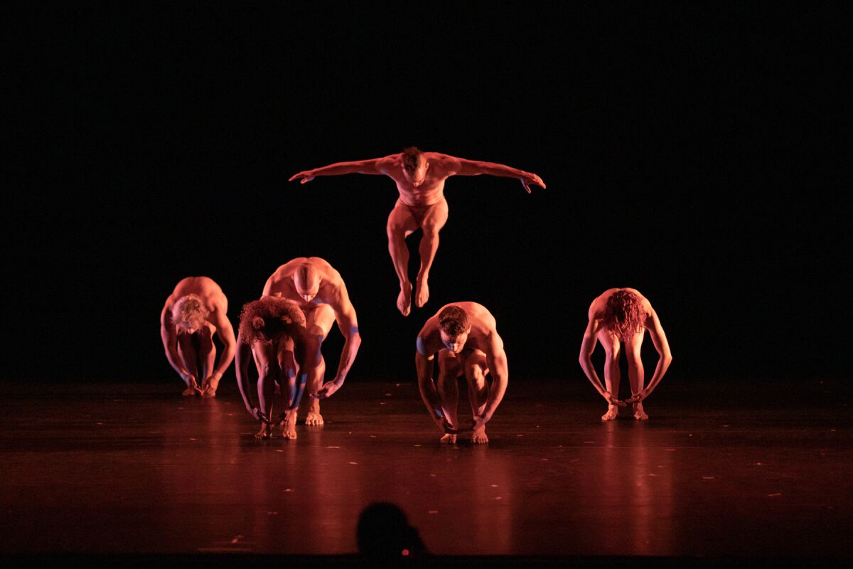 Five dancers hunched over while one in the middle jumps up with arms out.