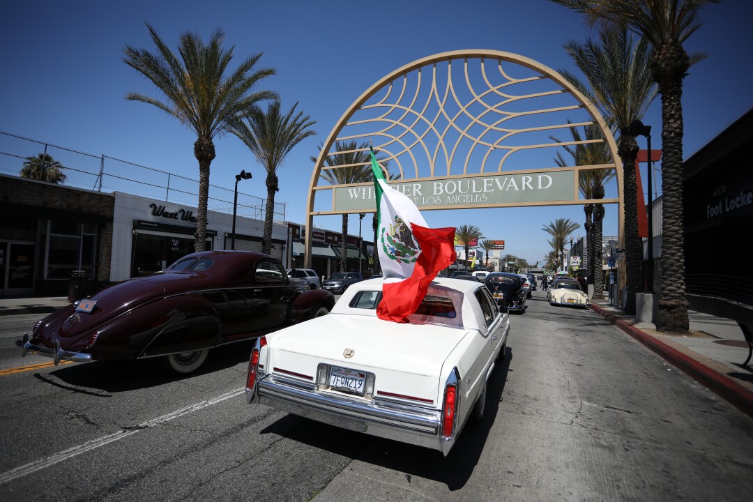 A Mexican flag billows from a car as it drives beneath a sign spanning a roadway that says "Whittier Boulevard."