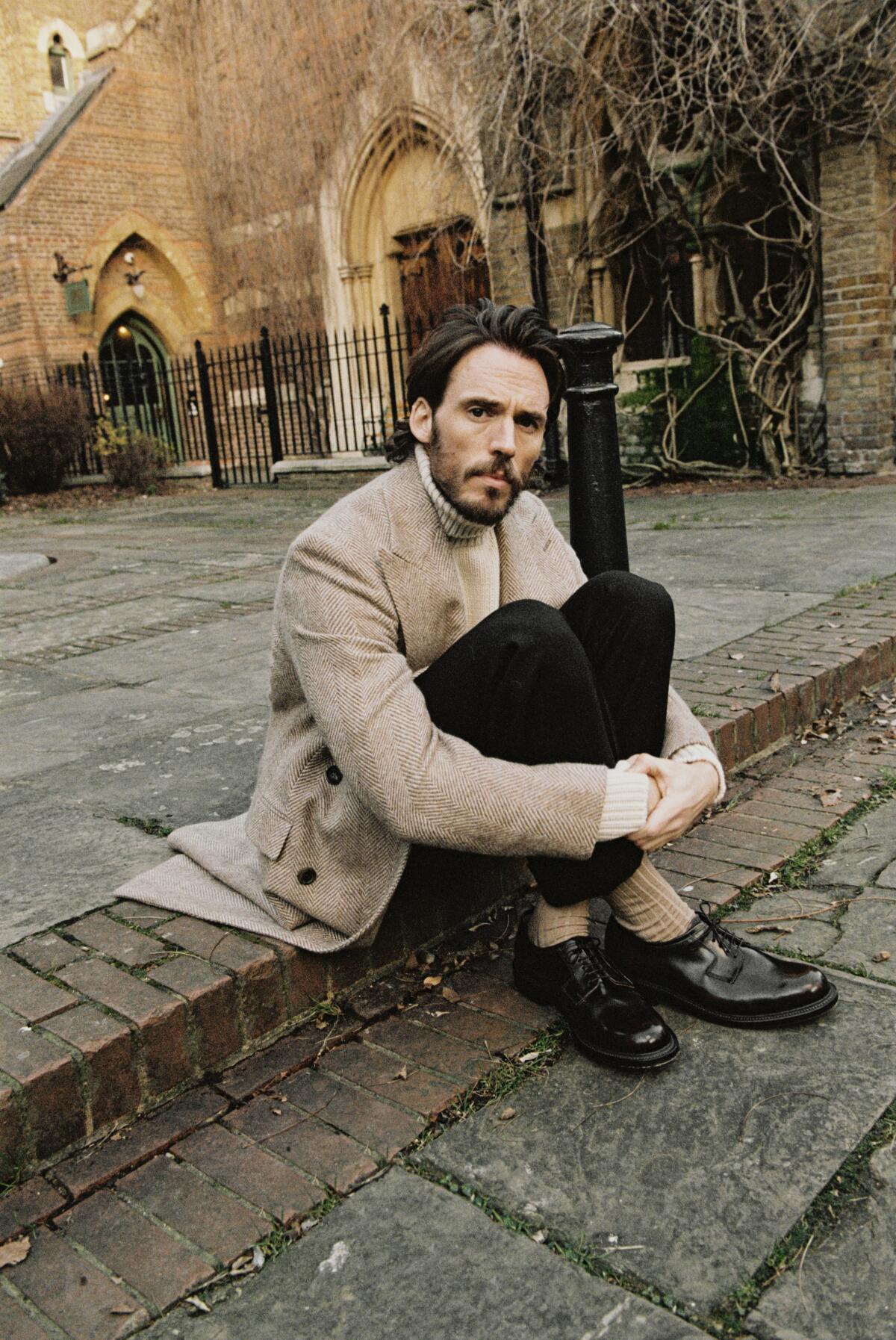 A bearded man (actor Sam Claflin) in a brown tweed jacket sits on a brick curb.