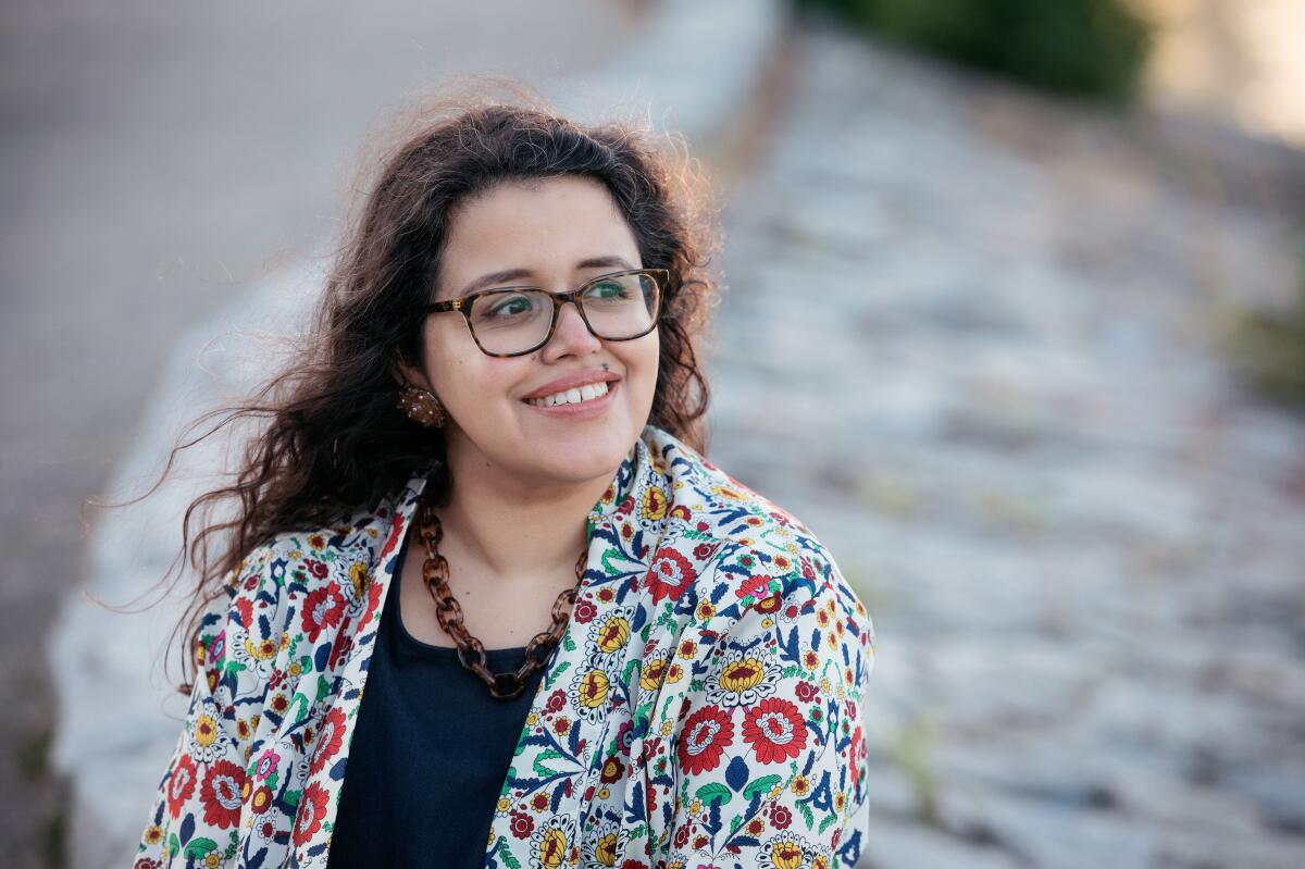 A smiling woman in glasses and a colorful blazer seated outdoor, looking off-camera