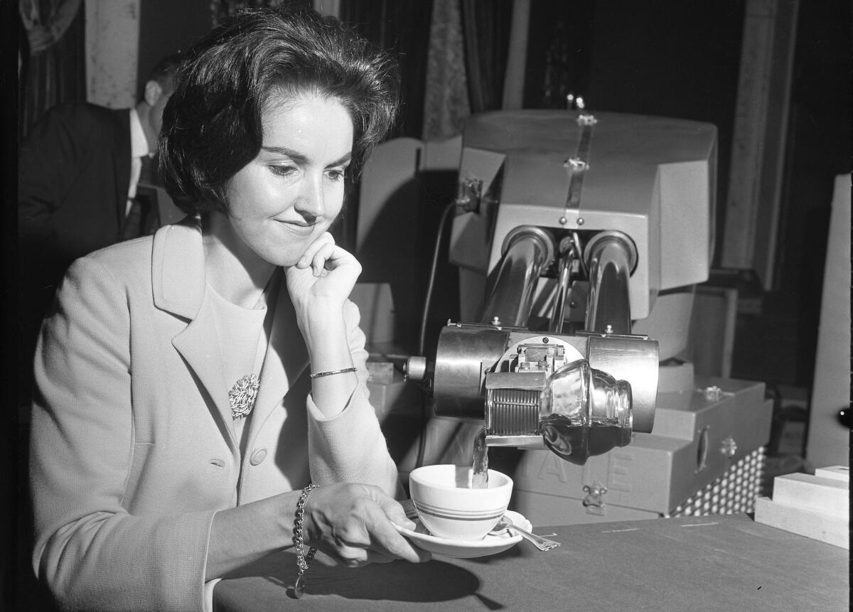 A black-and-white photo of a woman at a table holding a coffee cup as coffee is poured from a machine.