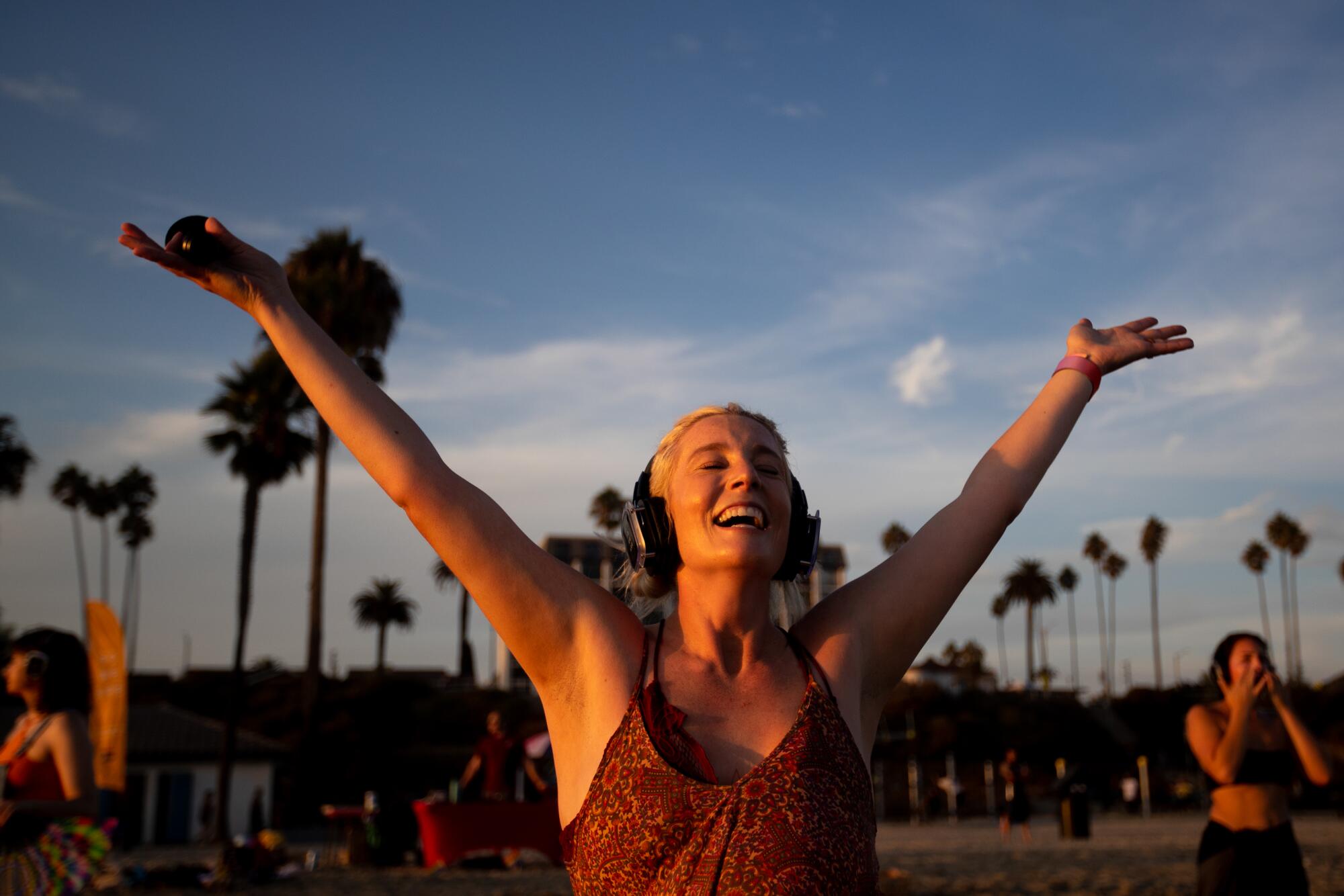 A woman wearing headphones with her arms raised into the air, on the beach at sunset.