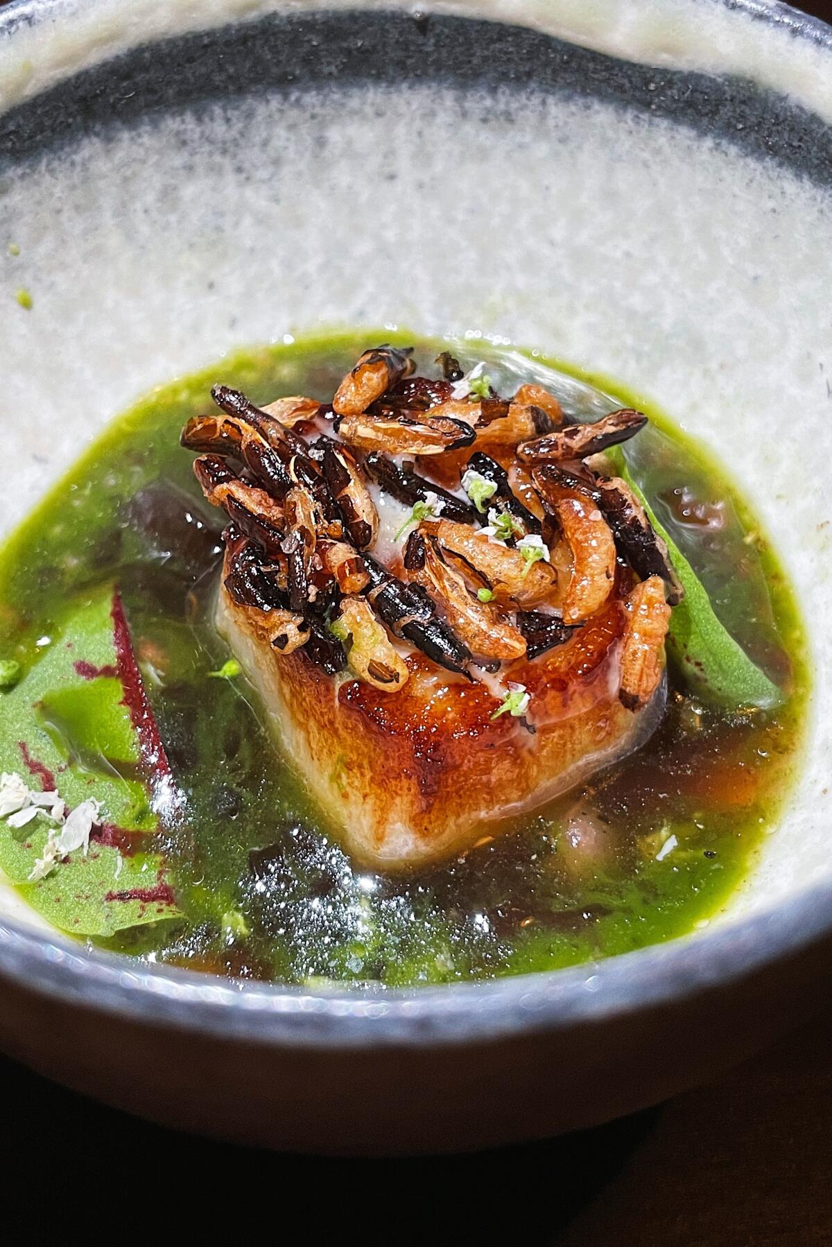 A browned Hokkaido scallop topped with puffed rice sits in green broth in a ceramic bowl at Baroo LA.