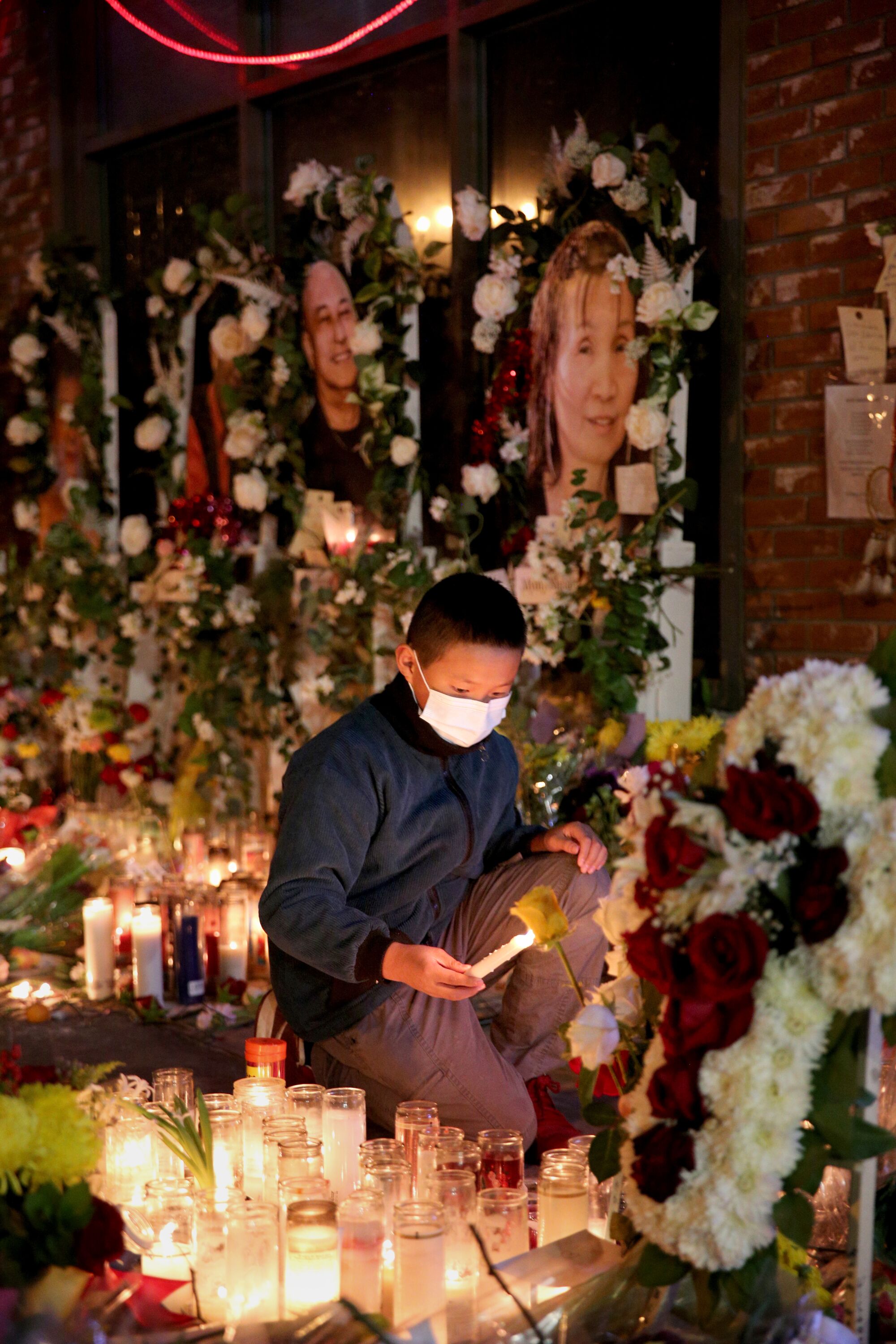 A boy lights candles at a memorial site , surrounded by lit candles and flowers, with portraits of victims above him.