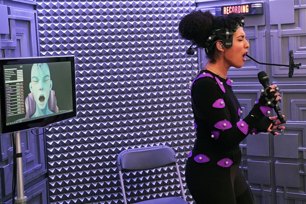 A singer wearing a bodysuit with sensors on it sings into a microphone in front of a monitor