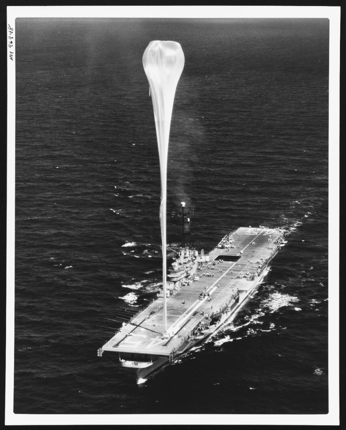 A very tall weather balloon moored to a U.S. aircraft carrier in an old black-and-white photo.
