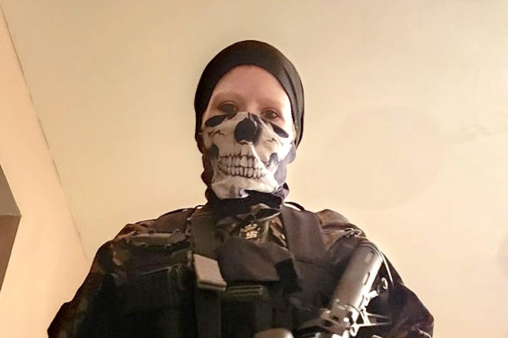 A woman with a skull bandanna around her face wears tactical gear while holding a rifle