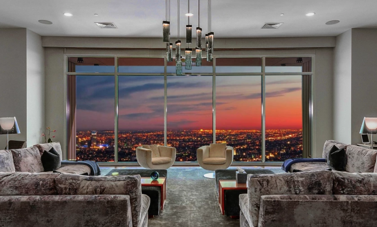 A floor-to-ceiling window in a modern-looking living room has a panoramic view of city lights at dusk.