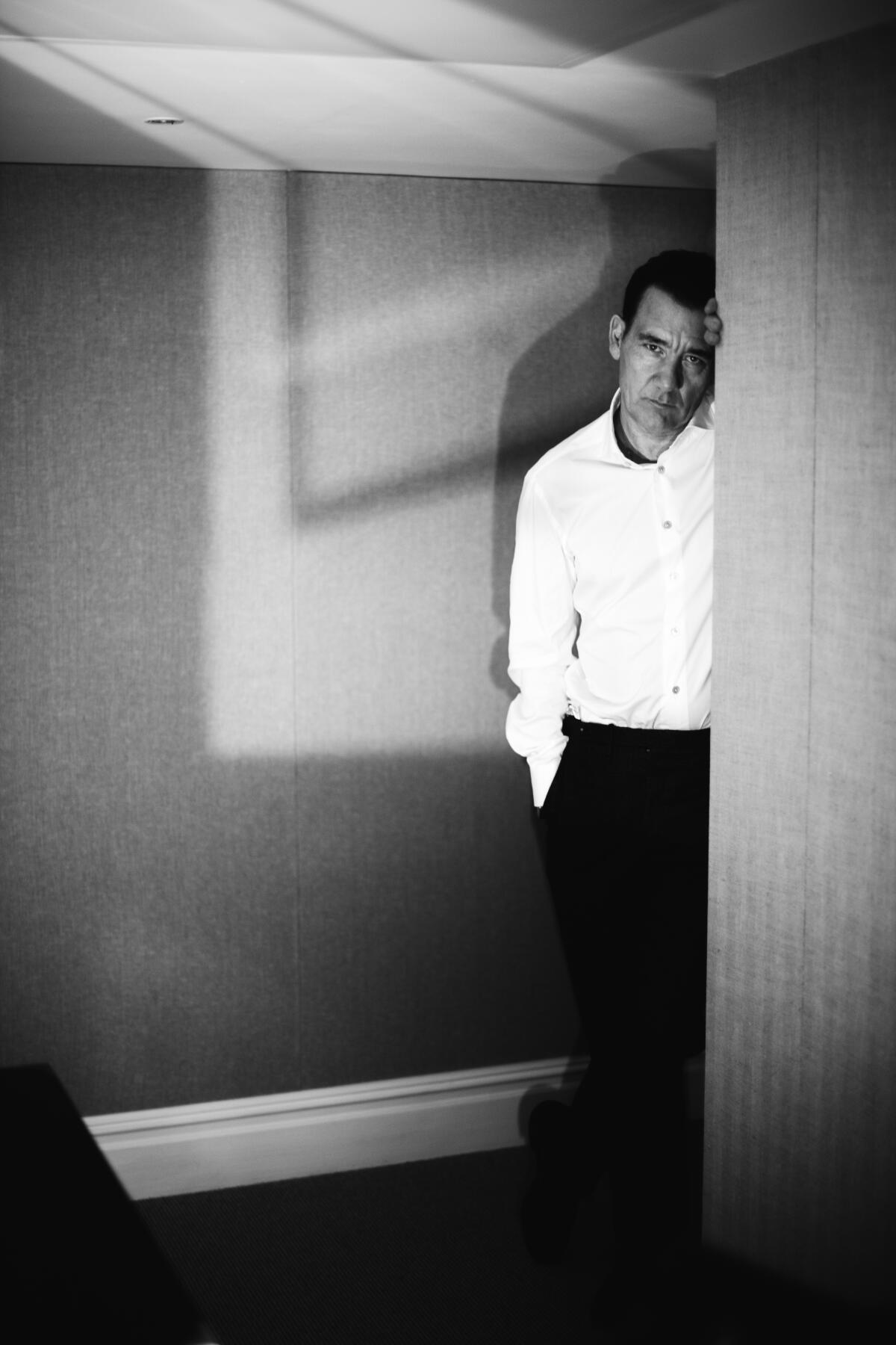 Clive Owen in a white shirt and dark pants leans his head against a door.