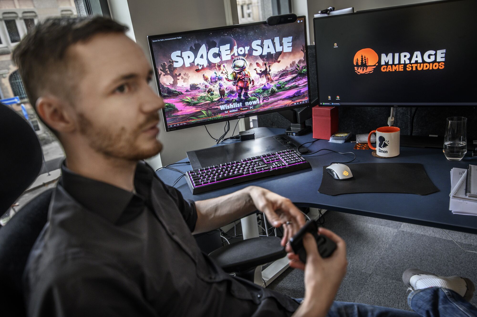 Un homme en chemise sombre assis à un bureau avec deux écrans d'ordinateur ; l'un dit Space for Sale, l'autre Mirage Game Studios