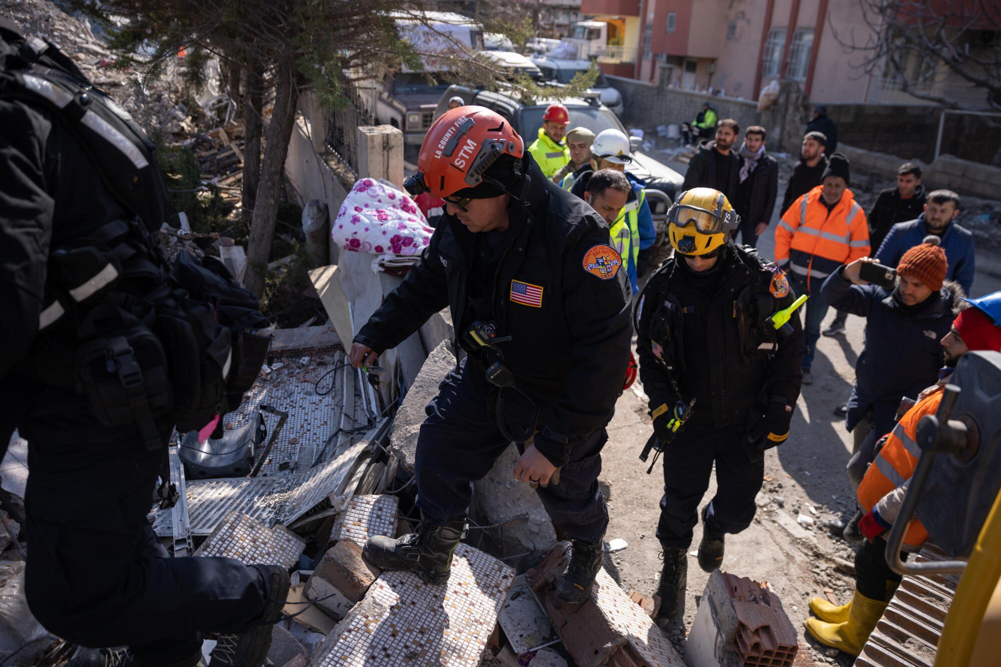 People in hard hats step over rubble amid buildings in the daytime.