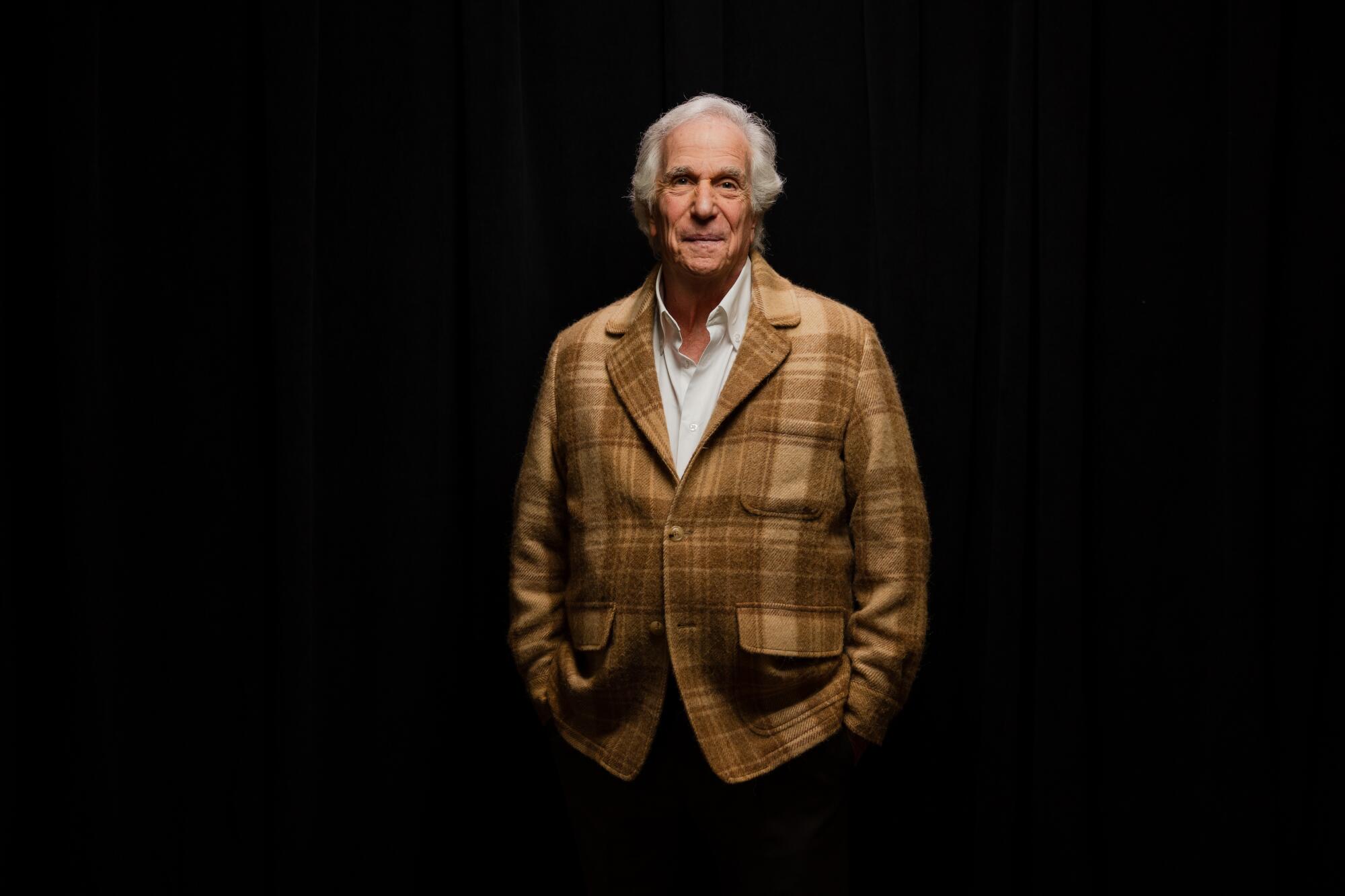 A man in a brown plaid jacket stands against a black background.