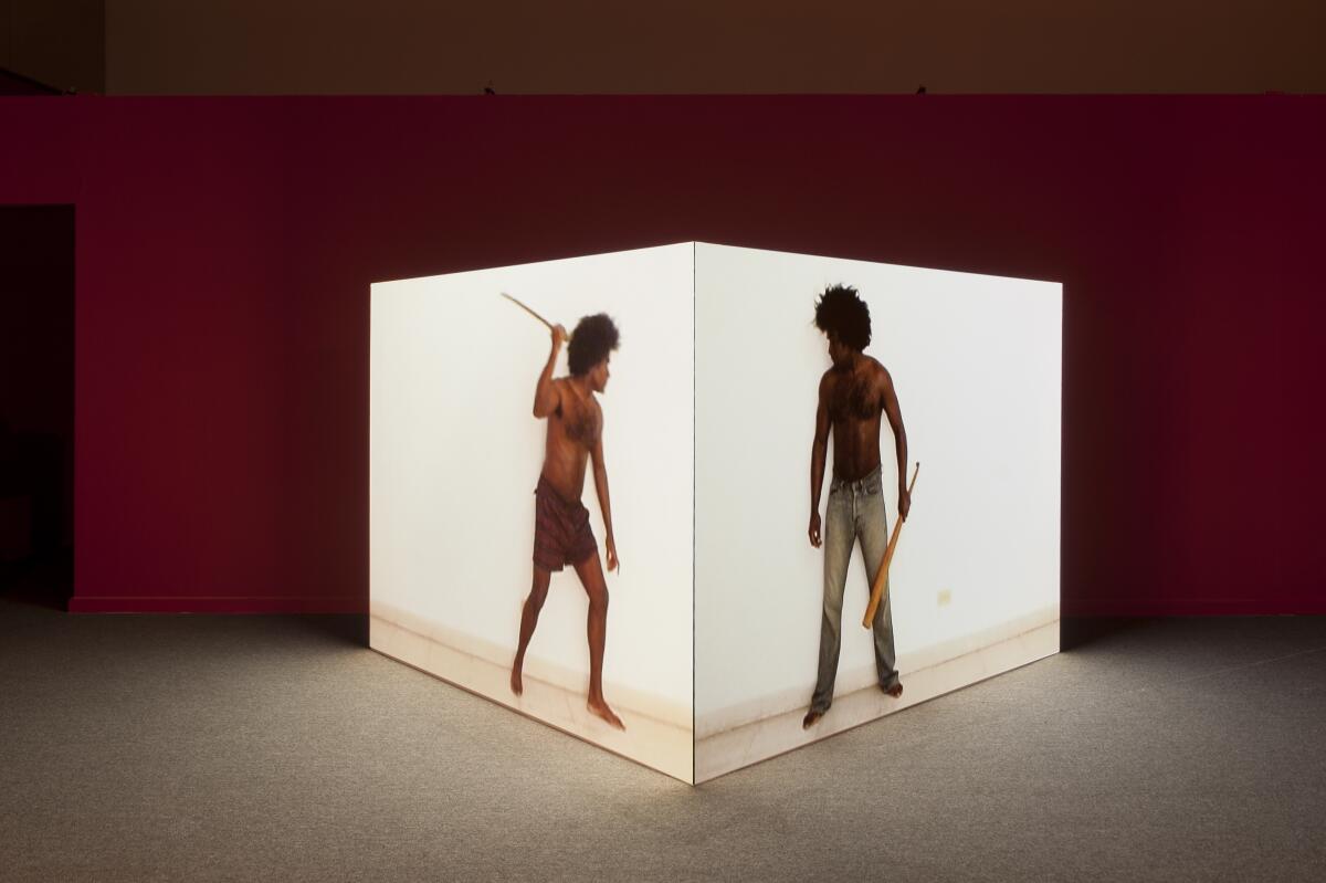A video work projected on a cube shows two Black male figures (the same man) wielding sticks on different surfaces.