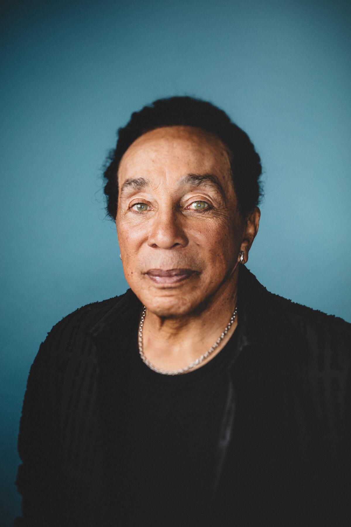 Smokey Robinson looks straight ahead in a black outfit with a gold chain and a serious expression