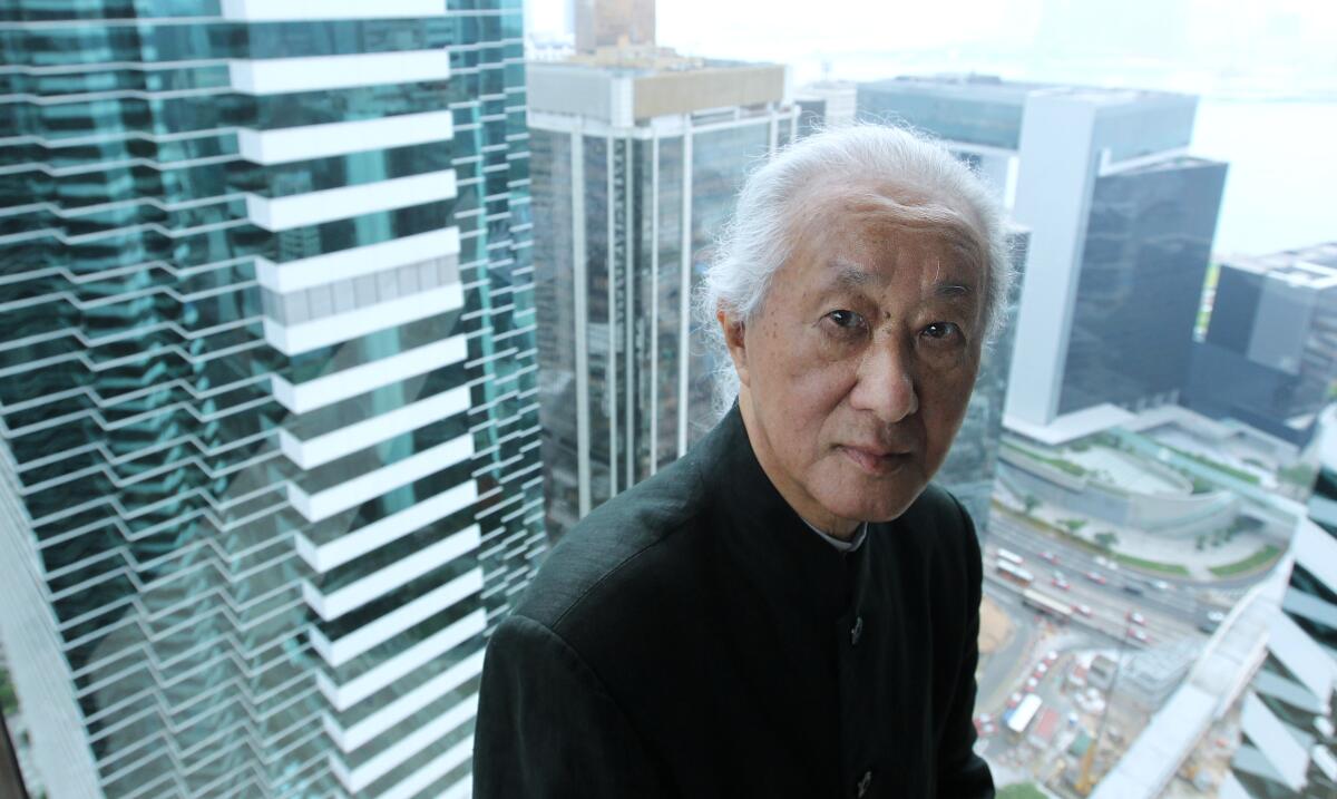 Arata Isozaki is seen sitting before a window with a view of a city behind him