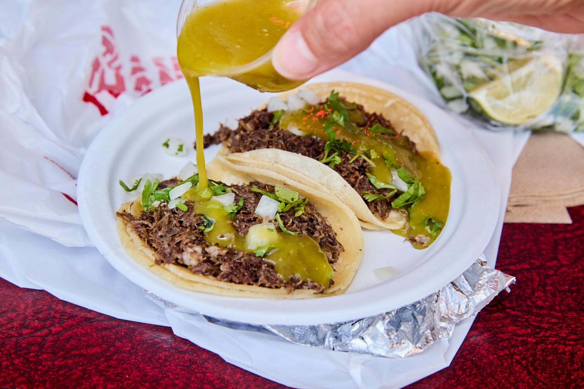 A hand pours green salsa from a plastic cup onto two tacos on a plate.