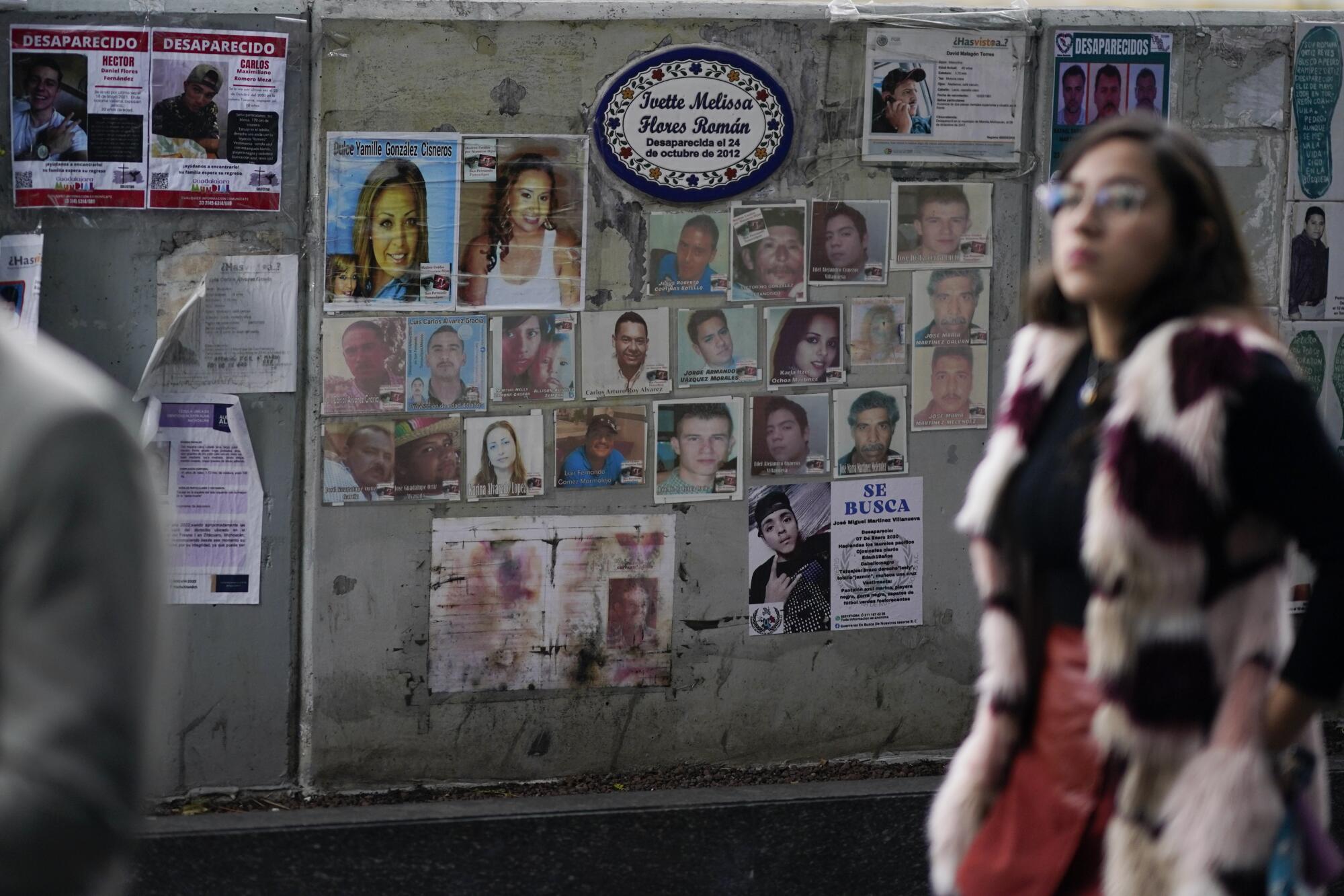 A woman with dark hair and wearing glasses walks near a wall bearing photographs of individual people 