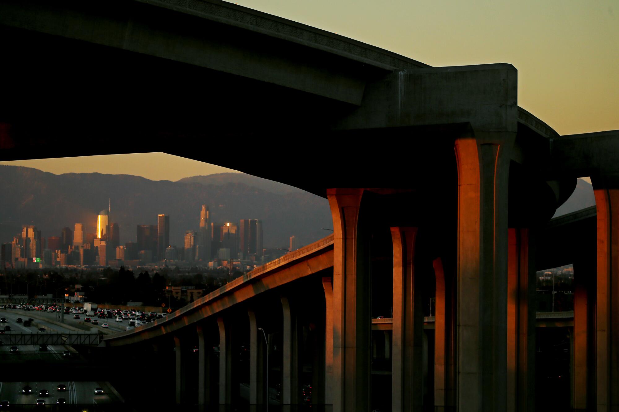 Smog hangs in the air as the sun sets on an elevated freeway, with the Los Angeles skyline in the distance. 