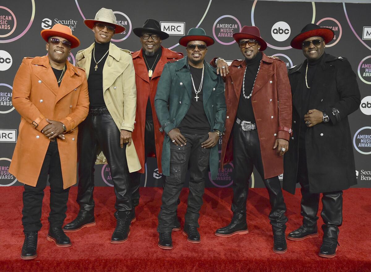 Six men wearing fedora hats and colorful long coats over black clothing 