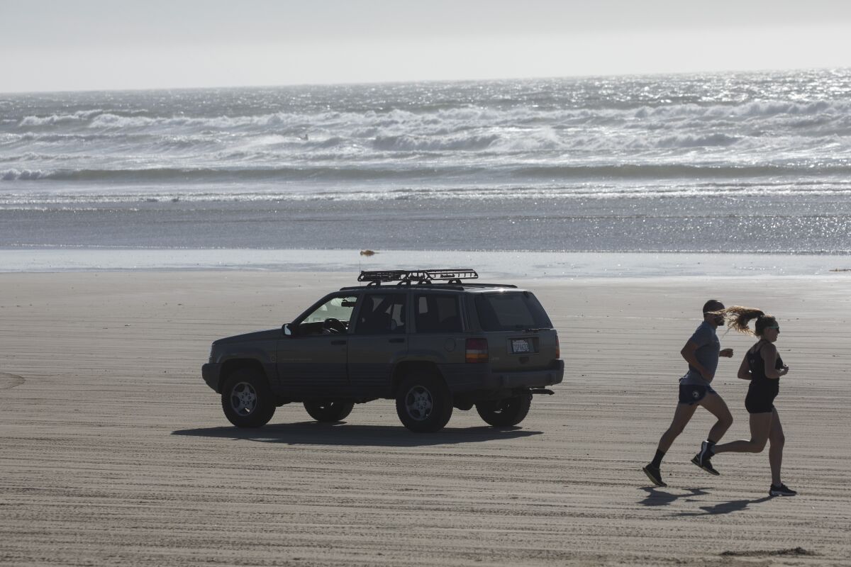 An SUV drives on a sandy beach as a couple jogs in the opposite direction.