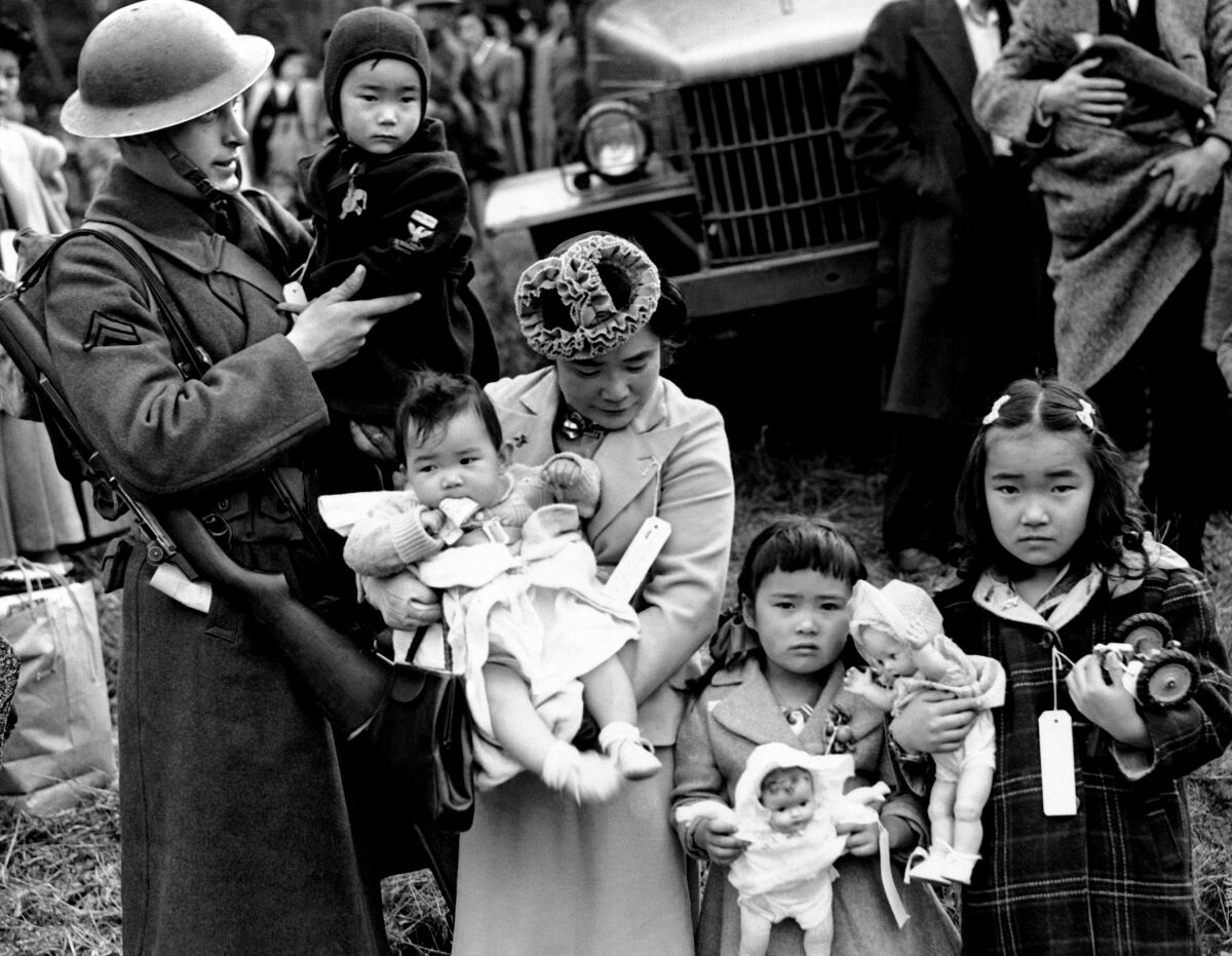 A man in military gear with a gun holds a baby next to a woman holding a baby and two small girls with dolls.