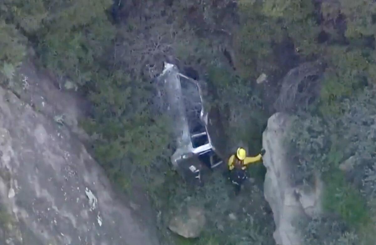 As seen from above, a car is smashed in a brushy area at the bottom of a cliff. A firefighter in yellow stands alongside.
