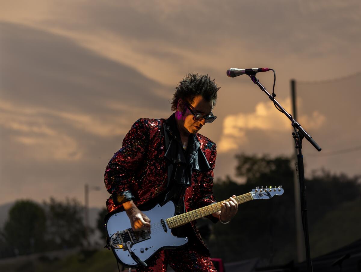 Musician Daniel Ash in sunglasses playing an electric guitar against a cloudy sky