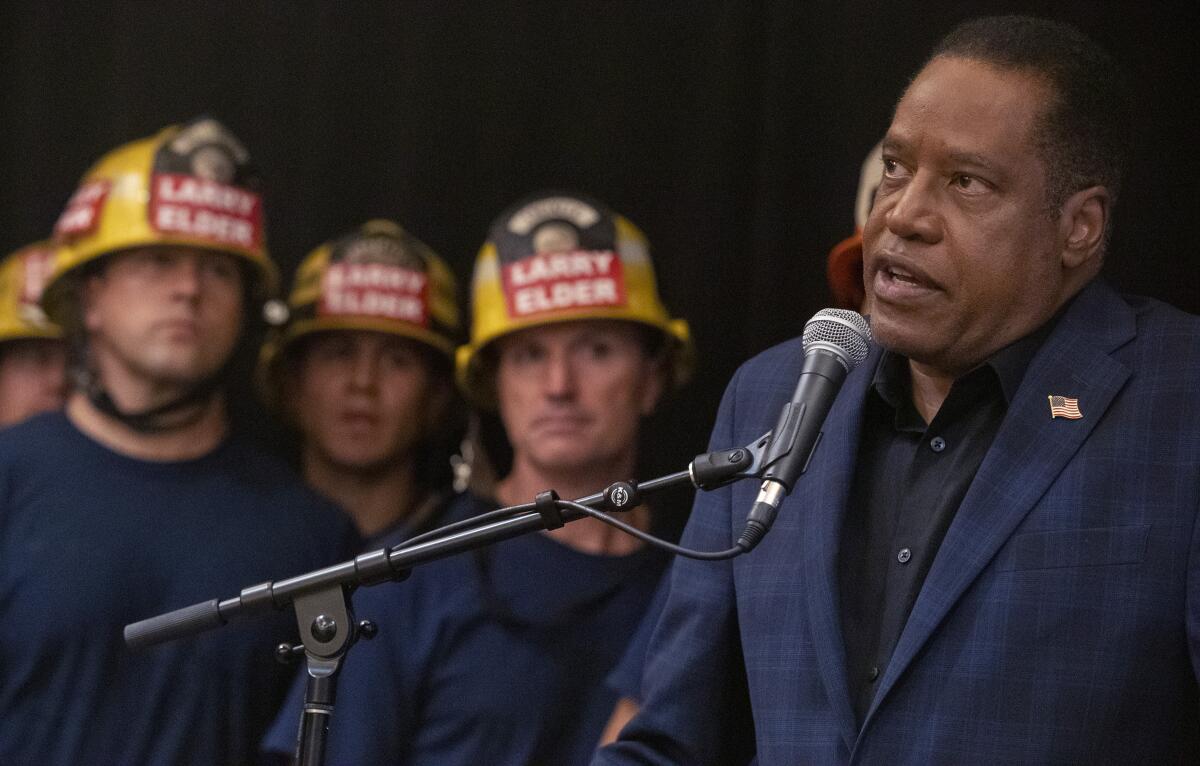 Larry Elder speaks into a microphone next to firefighters with "Larry Elder" helmets 