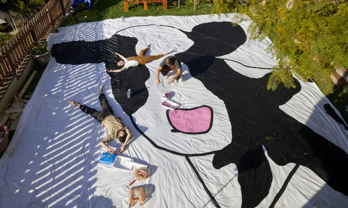 Three men lie on and paint a large sheet with a picture of a cow.