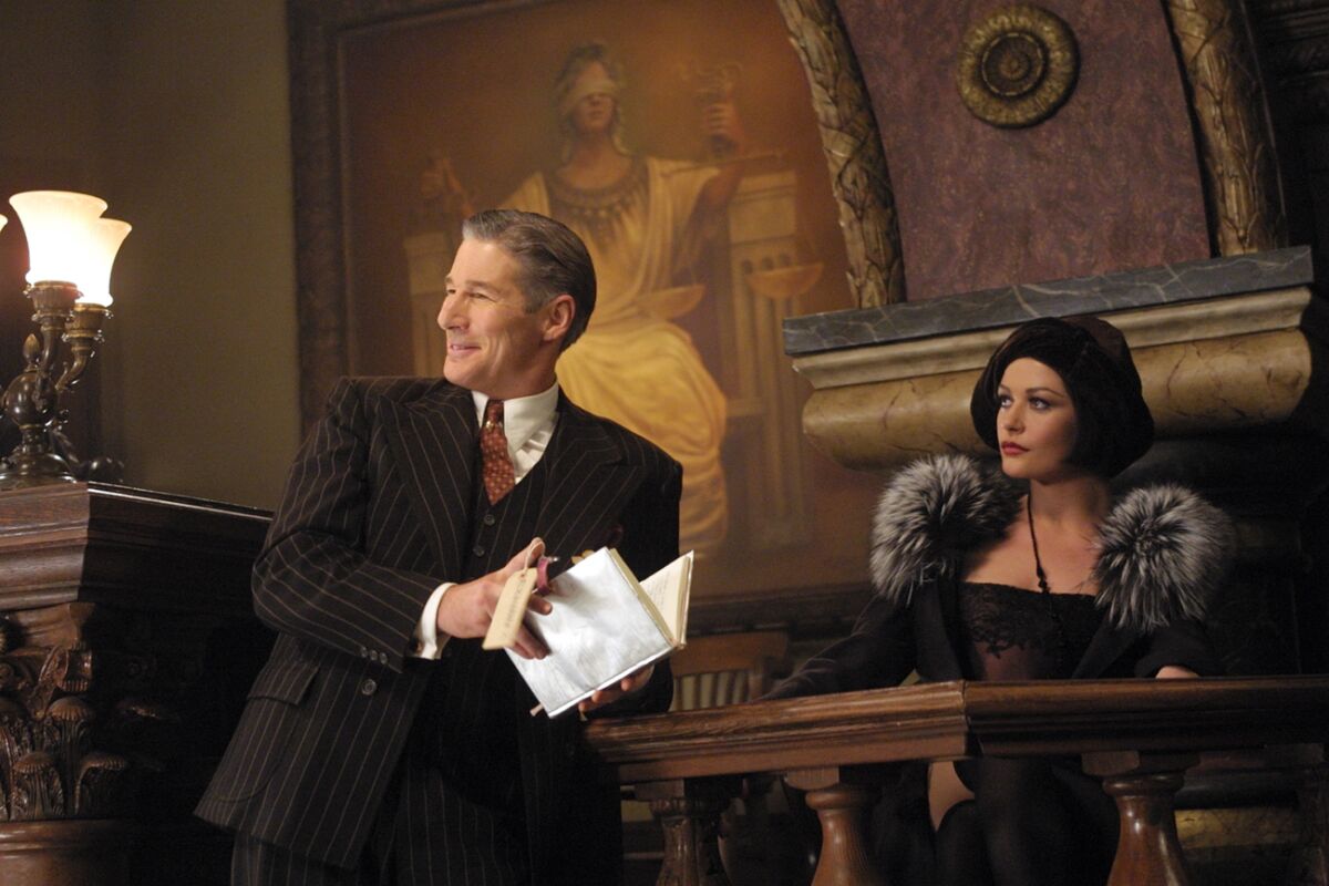 Richard Gere smiles while looking to the side as Catherine Zeta–Jones sits in the witness stand in “Chicago.”