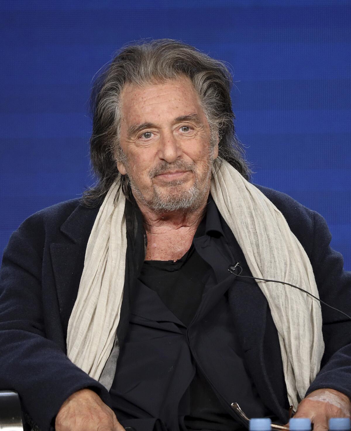 Al Pacino is wearing a black coat with a beige scarf as he smiles and sits on a stage.