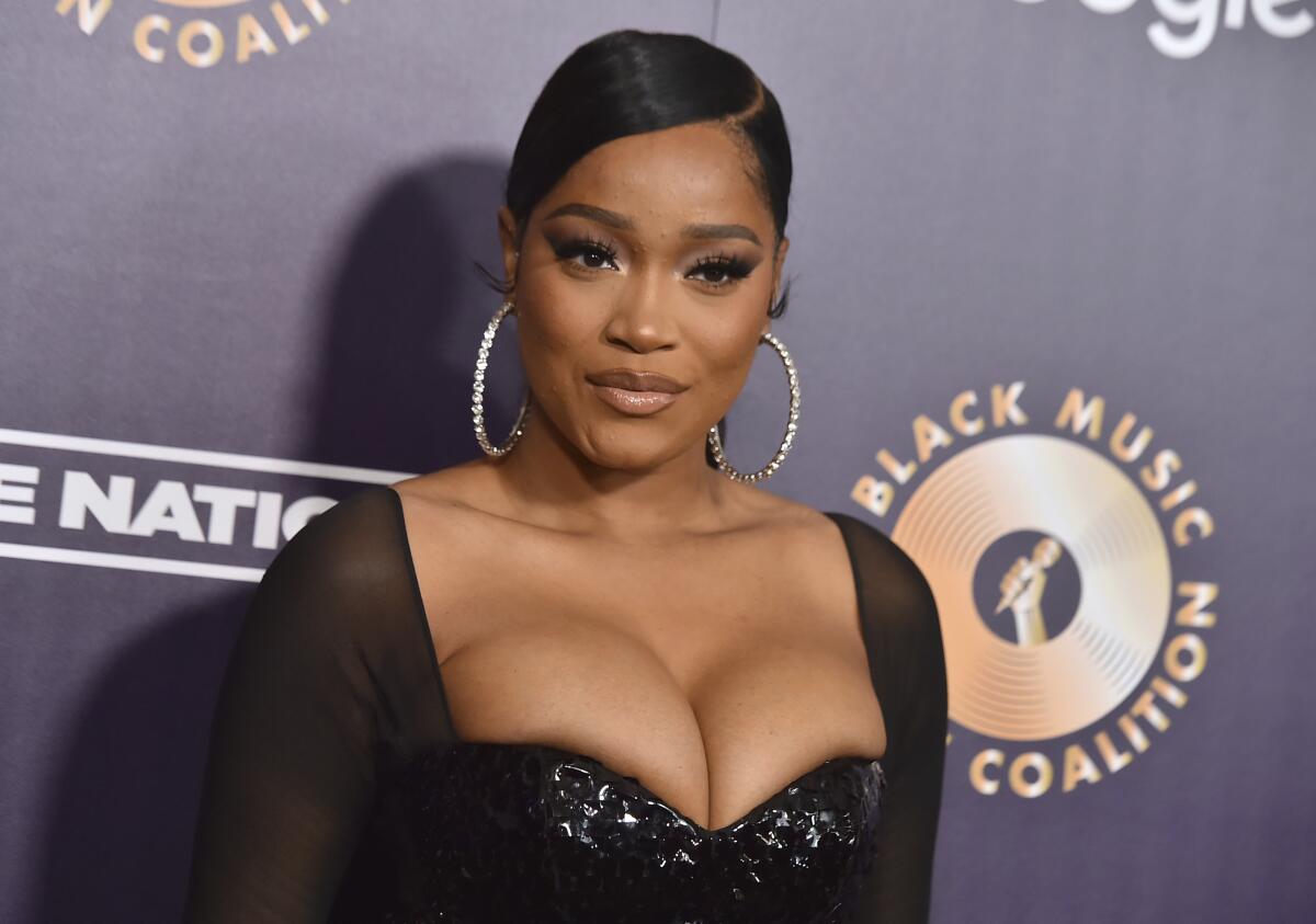 Keke Palmer poses with a serious face and slicked-back hair, wearing a low-cut black gown and large hoop earrings