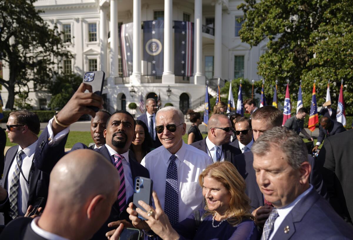 President Biden, wearing aviator sunglasses, poses for a photo amid a group of people on the South Lawn of the White House.
