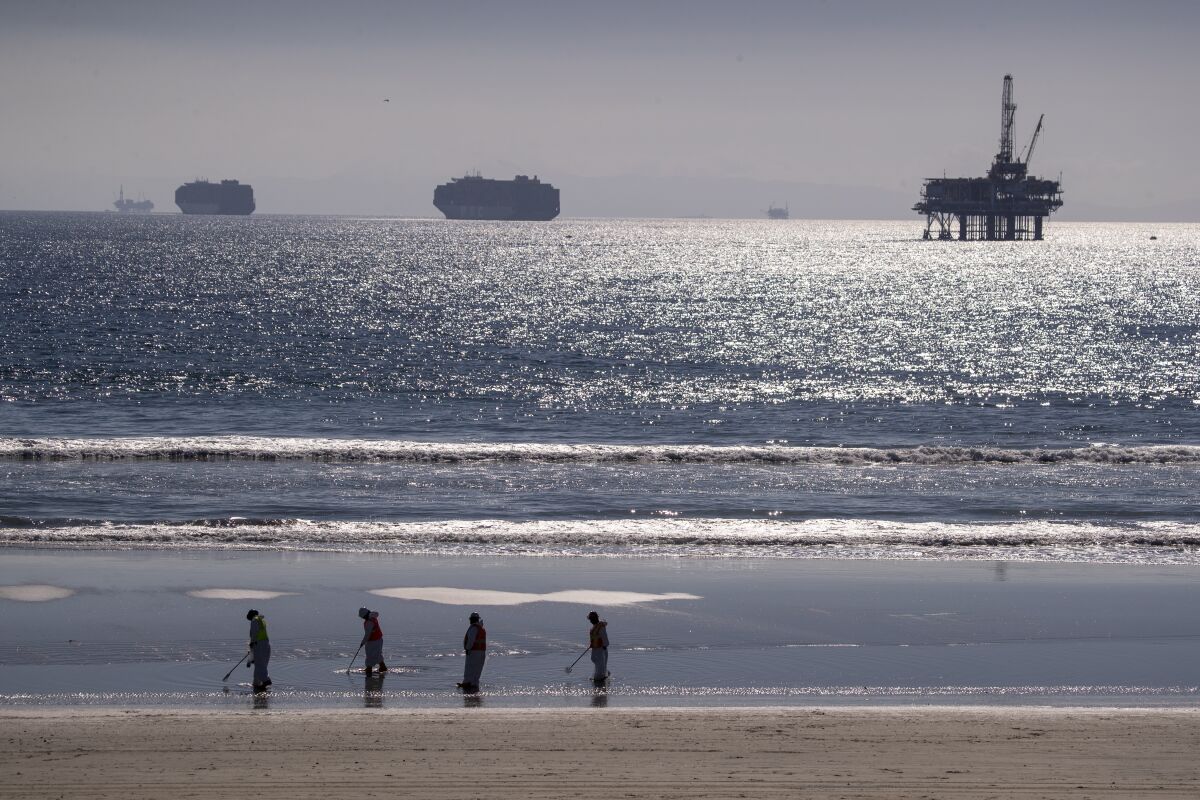 Container ships and an oil derrick line the horizon as environmental oil spill cleanup crews search the beach