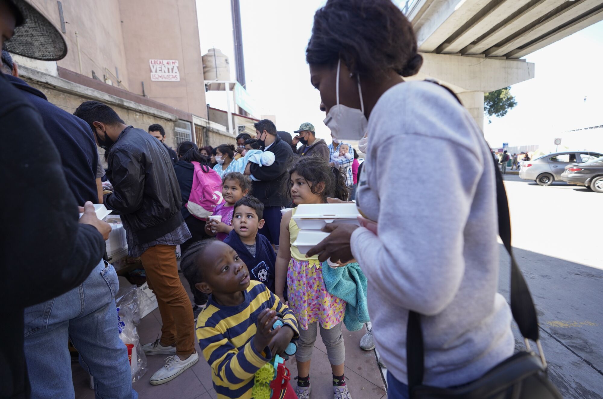Migrants, including small children, wait in line for food.