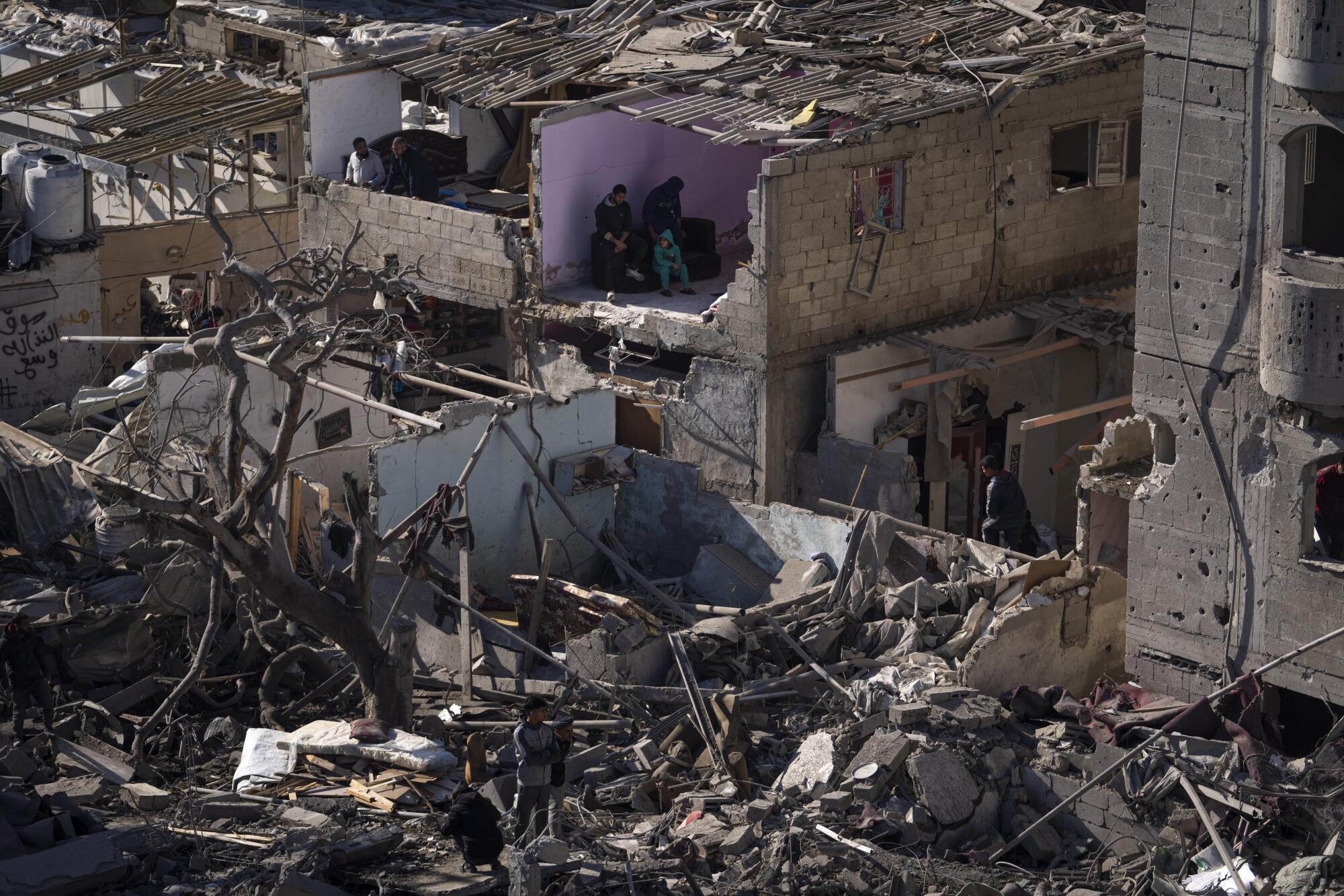 People sitting in what's left of a room as others survey the rubble of bombed-out buildings