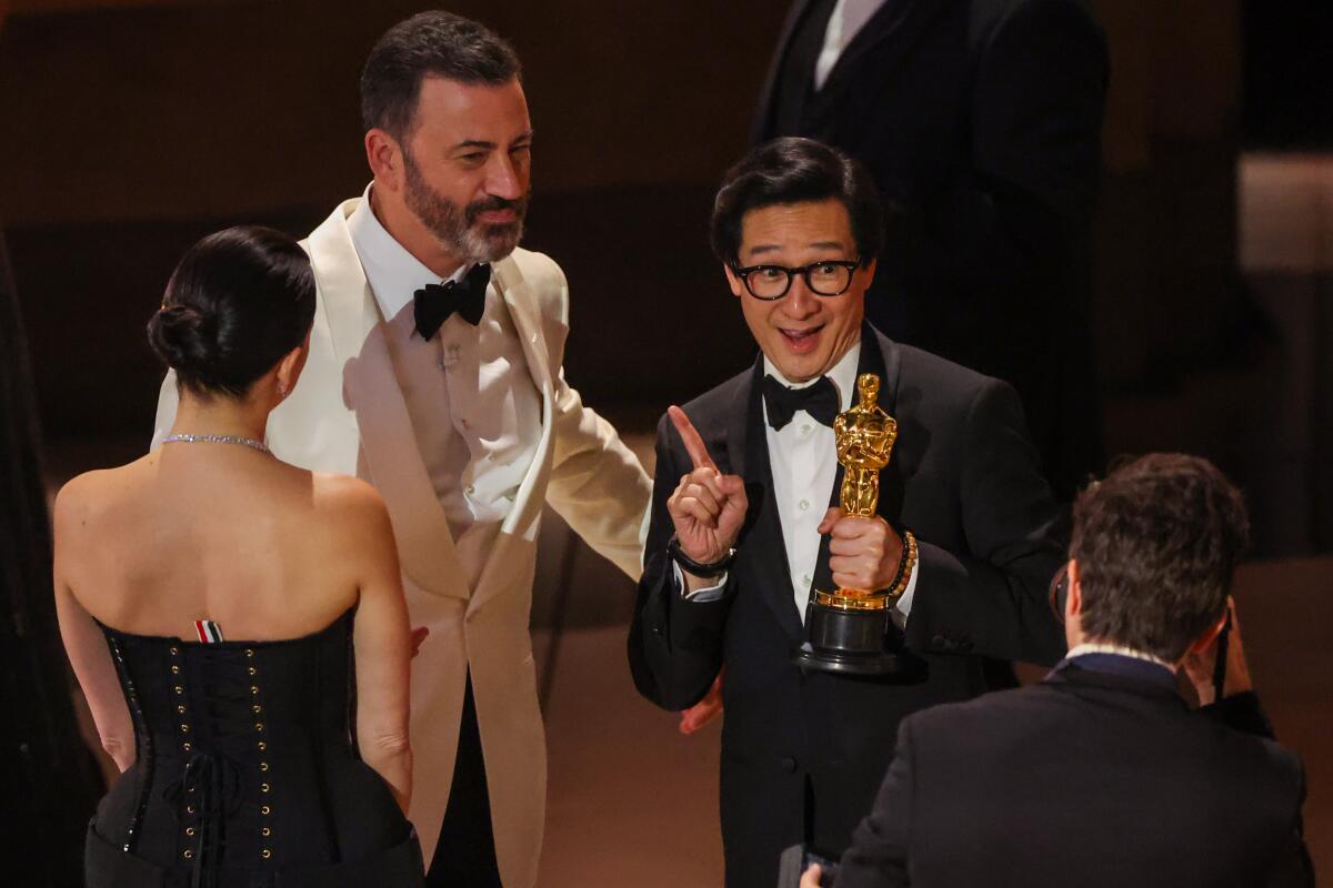 Jimmy Kimmel, in a white tuxedo, looks at Ke Huy Quan, in a black tuxedo, who is holding an Oscar.