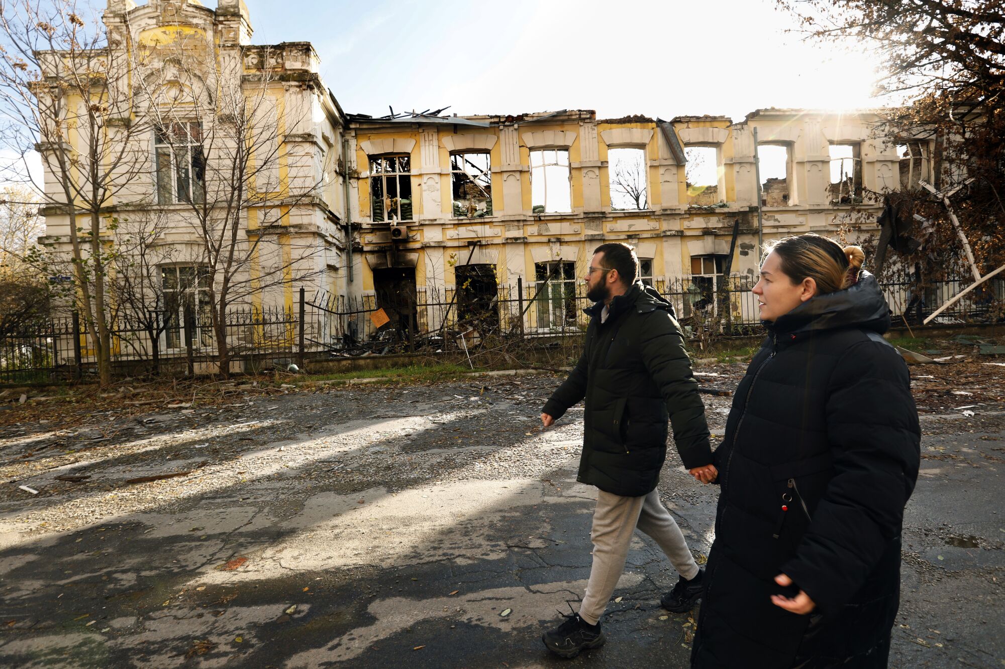 A bearded man, left, and a woman, both in dark winter jackets, hold hands as they walk past a destroyed building